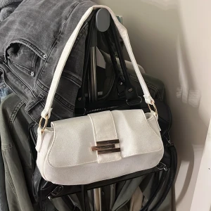 Vit axelväska - Säljer en stilren vit axelväska från ASOS i bomullstyg. Väskan har en avlång form, bred axelrem och snyggt guldfärgat spänne framtill. Perfekt för att lyfta din outfit med en clean och modern touch. Pris går att diskutera🤍