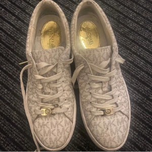 Beige sneakers från Michael Kors - Snygga beige sneakers från Michael Kors med MK-logga i mönstret och gulddetaljer på snörningen. Skorna har rund tå, platt sula och är tillverkade i textilmaterial med tydlig logotyp på sidan av sulan. Perfekta för dig som gillar stilrena och trendiga skor.