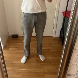 Grå raka jeans i bomull - Säljer ett par grå jeans med rak passform och klassisk femficksdesign. Jeansen är tillverkade i bomull och har en enkel, stilren look som passar till det mesta. Färgen är ljusgrå och materialet känns mjukt och följsamt. Storlek 30-32. På sista bilden är ett litet hål i samband med de nöttna. Hålet är lika stor som en ärta max