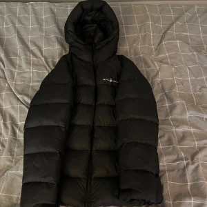 Svart pufferjacka från Sail Racing - Säljer en svart pufferjacka från Sail Racing med stor huva och dragkedja framtill. Jackan har en clean look med logga på bröstet och en stor vit detalj på huvan. Perfekt för kalla dagar när du vill hålla stilen och värmen. Hör av er vid frågor eller om ni vill se fler bilder😜👍