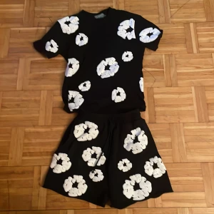 denim tears tracksuit - Svart t-shirt från Denim Tears med stora vita blommiga tryck över hela plagget. Klassisk passform med rund hals och korta ärmar. Materialet känns mjukt och skönt, perfekt för en chill och trendig look.