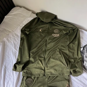 Olivgrön Jordan PSG Träningsoverall - Stiligt PSG x Jordan-set i olivgrönt med guldiga detaljer. Består av hoodie med dragkedja och matchande byxor. ✔️ Storlek: XL (158–170 cm, motsvarar ca S i vuxen) ✔️ Färg: olivgrön med guldiga loggor (PSG, Jordan, Qatar Airways) ✔️ Skick: Mycket bra, sparsamt använd. ✔️ Material: bekvämt och stretchigt tyg, perfekt för både träning och vardag.  En eftertraktad tracksuit för PSG- och Jordan-fans!  📦 Skickas snabbt och välpackat, eller möts upp vid intresse.