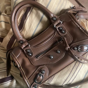Brun axelväska i skinnstil - Cool brun axelväska med både handtag och lång axelrem. Väskan har snygga metalldetaljer, dragkedja framtill och dekorativa spännen. Perfekt storlek för det viktigaste och ger en edgy vibe till din outfit.