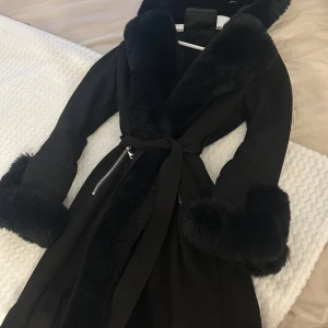 Svart kappa med fuskpäls och bälte - Snygg svart kappa med bred fuskpäls runt krage och ärmslut. Jackan har knytbälte i midjan och dragkedja i sidan. Perfekt för kalla dagar när du vill vara både varm och stilren.