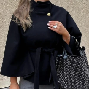 Poncho kappa - Stilren svart polotröja med hög hals, dekorativ guldig knapp vid axeln och knytbälte i midjan. Tröjan har vida ärmar och en loose passform som ger en cool och modern look. Perfekt för dig som gillar minimalistisk stil.