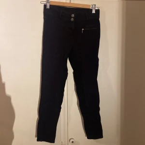 Svarta ridbyxor från Milton, stl 150 - Svarta ridbyxor från Milton med snygg passform och dragkedjeficka framtill. Byxorna är tillverkade i stretchigt material med 95% bomull och 5% elastan för extra komfort. Perfekta för ridning och har förstärkt säte för bättre grepp i sadeln. Använda sparsamt samt utan defekter eller synliga tecken på användning 