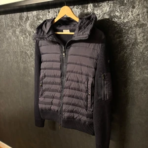 Mörkblå Moncler Cardigan - Snygg mörkblå cardigan från Moncler med stickade ärmar och huva. Jackan har quiltad front, dragkedja hela vägen och praktiska fickor med knappar. Ärmarna är ribbstickade och det finns en extra ficka med dragkedja på ena ärmen. Perfekt nu till hösten. 