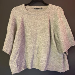 Grå stickad tröja från 157 - Mysig grå stickad tröja från 157 med rund halsringning och kortare ärmar. Tröjan har en loose passform och är perfekt för lager-på-lager. Enkel och stilren design som funkar till många olika outfits.