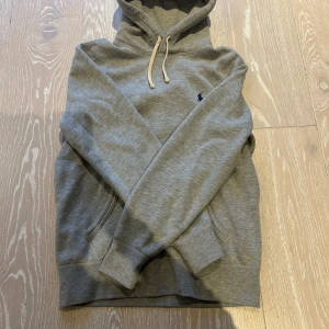 Grå hoodie från Polo Ralph Lauren - Säljer en grå hoodie från Polo Ralph Lauren med klassisk broderad logga på bröstet. Tröjan har huva med snörning, känguruficka och är tillverkad i mjuk bomullsblandning. Perfekt för chill dagar eller när du vill ha en clean och stilren look.