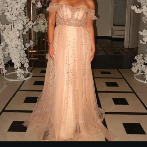 Korallrosa/champange färgad offshoulder balklänning - Drömmig långklänning i korallrosa/champagnefärg med offshoulder och glittriga detaljer. Klänningen har ett flowigt tylltyg, markerad midja och vackra paljetter som ger extra lyxig känsla. Perfekt för bal eller speciella tillfällen.