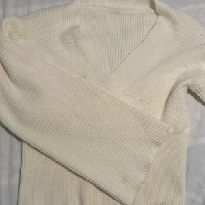 Ribbad vit omlott-tröja - Ribbad vit omlott-tröja med v-ringning och långa ärmar. Tröjan är stickad och har en mjuk känsla, perfekt för lager-på-lager under kyligare dagar. Enkel och stilren design som passar till det mesta.