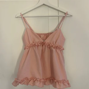 Ljusrosa volanglinne med tunna axelband - Supersöt ljusrosa linnetopp med tunna axelband och volangdetaljer både under bysten och längst ner. Toppen har en lätt strukturerad yta och är luftig, perfekt för varma dagar. Passformen är loose och toppen är ärmlös.