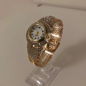 Glittrig guldig damklocka med stenar - Säljer en riktigt snygg damklocka från Quartz med glittrande stenar på armbandet och runt urtavlan. Klockan har en rund vit urtavla med svarta siffror och guldfärgade visare. Armbandet är guldigt och har ett unikt spiralformat mönster.                                          ✅Frakt inom 24 timmar 📦.                                                         ✅Nytt batteri 🔋.    