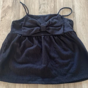 Svart linnetopp med rosett från Kappahl - Supersöt svart linnetopp från Kappahl med smala axelband och en stor rosett framtill. Toppen är tillverkad i ett lätt, krinklat tyg som ger en cool struktur. Perfekt för dig som gillar en enkel men ändå söt look.