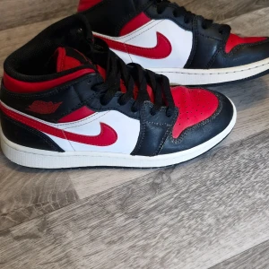 Nike Air Jordan 1 Mid Svart/Röd/Vit - Säljer ett par Nike Air Jordan 1 Mid med klassisk färgblockning i svart, rött och vitt. Skorna har svart skinn, röda detaljer och vit mellansula. Ikonisk röd swoosh och Jordan Wings-logga på sidan. Perfekta för dig som vill sticka ut med en tidlös sneaker.