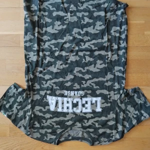 Camouflage t-shirt från Lechia Gdansk - Säljer en grön och grå camouflage t-shirt från Lechia Gdansk med tryck på bröstet. T-shirten har korta ärmar och rund hals. Perfekt för dig som gillar sportiga och street-inspirerade plagg.