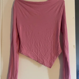 Rosa långärmad topp med asymmetrisk fåll - Säljer en rosa långärmad topp med unik asymmetrisk fåll som ger en cool och trendig look. Toppen har en enkel design utan mönster och är tillverkad i mjuk bomull. Perfekt för dig som vill sticka ut med en snygg och annorlunda siluett.