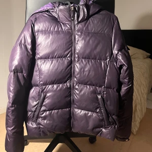 Lila pufferjacka från Champion S - Säljer en lila pufferjacka från Champion med huva och dragkedja framtill. Jackan har två fickor med dragkedja och en patch på ärmen. Materialet är glansigt och känns mjukt, perfekt för kalla dagar.