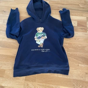 Mörkblå Polo Bear hoodie från Ralph Lauren - Mörkblå hoodie från Polo Ralph Lauren med ikoniskt Polo Bear-tryck på bröstet. Tröjan har huva, långa ärmar och en klassisk relaxed passform. Nypris ca 1500. Pris kan diskuteras, hör av er vid minsta fundering!