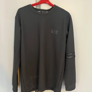 Grå långärmad tröja från EA7 Armani - Snygg grå långärmad tröja från EA7 Emporio Armani med rund halsringning. Diskret logga på bröstet och två svarta band med Emporio Armani-tryck runt ena ärmen. Perfekt för en stilren och sportig look.