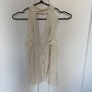 Krämvit volangtopp från Zara XS - Säljer en krämvit ärmlös topp från Zara i storlek XS. Toppen har djup v-ringning, volangdetaljer längs framsidan och en hög krage med knäppning i nacken. Lätt och flowigt material som ger en snygg siluett.