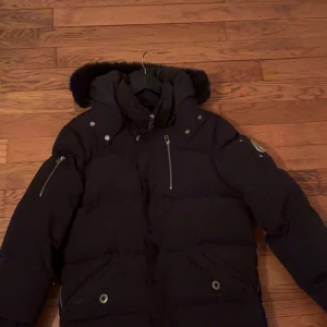 Svart dunjacka från Moose Knuckles - Säljer en svart dunjacka från Moose Knuckles med fluffig pälskant på huvan, flera dragkedjor och tryckknappar. Jackan har en cool patch på ärmen och är riktigt varm och skön för vintern. Perfekt för dig som vill ha en snygg och funktionell vinterjacka.