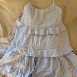 Ljusblå volangklänning från Nelly - Supersöt ljusblå klänning från Nelly med tunna axelband och flera lager volanger. Modellen är kort och har en luftig passform som ger en skön och chill vibe. Perfekt för varma dagar och har en feminin look med volangdetaljer. Materialet är lätt och känns mjukt mot huden. Aldrig använd, lappen sitter kvar 