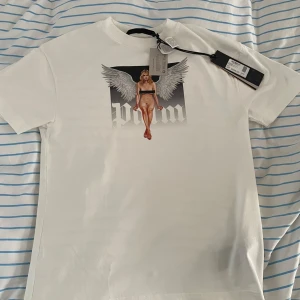 Palm Angels tshirt - (Passar också S) Snygg vit t-shirt från Palm Angels i 100% bomull. Framsidan har ett stort tryck med en ängel och texten 'Palm' i gotisk stil. Klassisk passform med korta ärmar och rund hals. Perfekt statement-plagg för dig som gillar streetwear vibes.