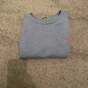 Ljusblå stickad tröja från Polo Ralph Lauren - Ljusblå stickad tröja från Polo Ralph Lauren med klassisk rund halsringning och broderad rosa logga på bröstet. Tröjan har långa ärmar och ribbade muddar vid ärmslut och nederkant. Perfekt för en chill och stilren look.