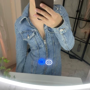 Kort jeansklänning med dragkedja - Säljer en snygg ljusblå jeansklänning med lång ärm och markerad midja. Klänningen har hel dragkedja framtill, klassisk skjortkrage och bröstfickor med knapp. Perfekt för dig som gillar denim och vill ha en cool, figurnära look.