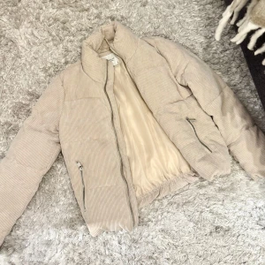 Beige manchester pufferjacka med dragkedja - Säljer en beige pufferjacka med mjuk manchester med diskreta ränder. Jackan har hög krage, två fickor med dragkedja och hel dragkedja framtill. Perfekt för kyliga dagar och ger en clean och trendig look.