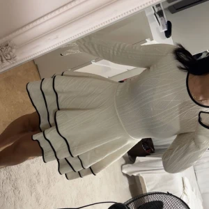 Vit kortklänning med svarta detaljer - Supersöt vit kortklänning med lång ärm och svarta kantdetaljer vid halsringning, ärmslut och volangkjol. Klänningen har ett diskret mönster och sitter snyggt med markerad midja och voluminös kjol. Perfekt för dig som gillar stilrena och trendiga plagg.