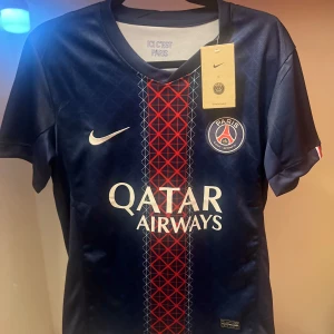PSG matchtröja Nike Dri-FIT blå/röd 25/26 - Paris Saint-Germain fotbollströja från Nike med Dri-FIT-material. Djupblå bas med röd och vit grafisk detalj i mitten, PSG-emblem på bröstet och vit Nike-logga. Kortärmad modell, perfekt för dig som älskar fotboll och vill sticka ut på planen.