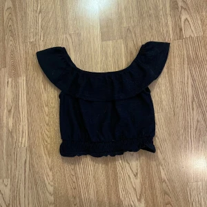 Svart offshoulder topp  - Svart croppad topp från River Island med bred volang runt axlarna och offshoulder-design. Resår i nederkant ger en snygg passform. Funkar som strl xxxs/xxs
