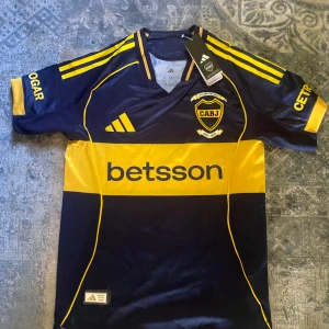 Boca Juniors matchtröja Adidas gul/blå - Snygg Boca Juniors fotbollströja från Adidas i mörkblått och gult med klubbmärke, sponsorer och klassiska Adidas-ränder på axlarna. Tröjan är kortärmad och tillverkad i ett lätt, ventilerande material perfekt för match eller träning.