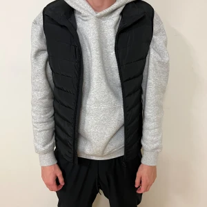 Svart dunväst med dragkedja - Säljer en svart dunväst med quiltad design och hel dragkedja framtill. Västen har en klassisk, enkel look och passar perfekt över en hoodie. Den är lätt och smidig, perfekt för lager-på-lager-stil.