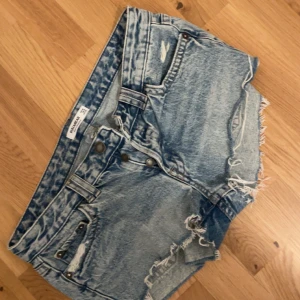 Blå jeansshorts från Pull&Bear - Snygga blå jeansshorts från Pull&Bear med låg midja och råa, fransiga kanter. Shortsen har slitna detaljer, klassiska fickor både fram och bak samt knappar i metall. Perfekta för sommarens alla äventyr och är korta men har en loose passform.