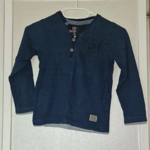 Blå långärmad tröja från S&R - Mörkblå långärmad tröja från S&R med smala ränder och tryck på bröstet. Tröjan har knappar vid halsen och en liten etikett nertill. Perfekt för dig som gillar en enkel men snygg stil.