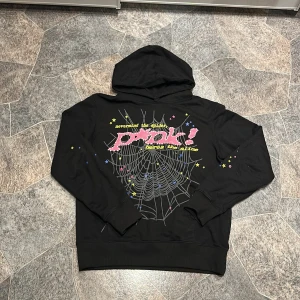Svart hoodie från Spåuer med tryck - Svart hoodie från Spåuer i 100% bomull med stor grafisk print på ryggen. Trycket har spindelnät, stjärnor och texten 'PUNK!' i rosa med vita prickar, samt gula och blå detaljer. Huva och muddar vid ärmslut och nederkant. Perfekt statement-plagg för dig som gillar unik streetstyle.