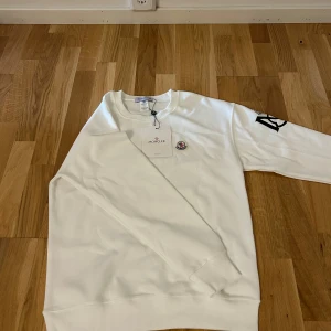Vit sweatshirt från Moncler med logga - Snygg vit sweatshirt från Moncler med klassisk rund halsringning. Tröjan har Moncler-logga på bröstet och en stor svart M-logga på ena ärmen. Tillverkad i mjuk bomull och har ribbade muddar vid ärmslut och nederkant. Perfekt för en clean och stilren look.