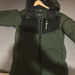 Grön pufferjacka från Sail Racing - Mörkgrön pufferjacka från Sail Racing med svarta detaljer på bröst och huva. Jackan har dragkedja framtill, två bröstfickor med dragkedja och en stor huva. Perfekt för kalla dagar och har en sportig vibe.
