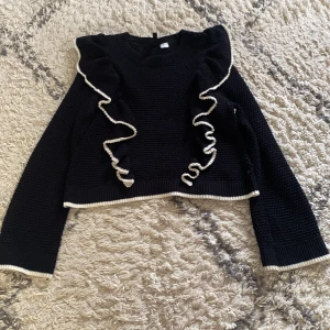 Svart stickad tröja med volangdetaljer - Supersöt svart stickad tröja med vita kanter och volangdetaljer framtill. Tröjan har lång ärm och är croppad i modellen. Perfekt för dig som vill ha något unikt och trendigt i garderoben!