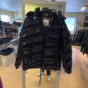 Svart moncler Maya 2 - Säljer en svart, glansig pufferjacka från Moncler med huva och dragkedja framtill. Jackan har Moncler-logga på ärmen, två fickor med dragkedja och justerbar dragsko nertill. Perfekt för kalla dagar och riktigt snygg streetstil.