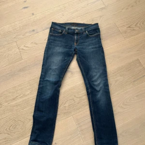  Nudie Jeans  - Snygga blå jeans från Nudie Jeans i storlek 29/32 med klassisk femficksdesign och kontrastsömmar. Modellen är tight long John och har Nudie Jeans signaturbroderi på bakfickorna. Perfekta för dig som gillar en smal siluett och stilren look.