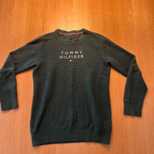 Mörkgrön stickad tröja Tommy Hilfiger - Mörkgrön stickad tröja från Tommy Hilfiger med broderad logga på bröstet. Klassisk rund halsringning och ribbade muddar vid ärmslut och nederkant. Perfekt för dig som gillar stilrena och snygga basplagg.