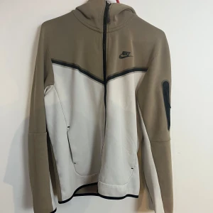 Nike hoodie i beige och vitt med huva - Nike hoodie i beige och vitt med svart logga på bröstet. Tröjan har hel dragkedja, huva och praktisk ficka med dragkedja på ena ärmen. Snygg kontrast mellan färgerna och sportig look. Perfekt för chill eller träning.
