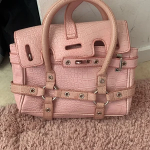 Rosa citybag  - Säljer en rosa handväska i croco-mönstrat syntetmaterial med coola silverfärgade nitar och ringdetaljer. Väskan har två rejäla handtag, spännen och dragkedja på insidan. Perfekt för dig som vill sticka ut med en edgy och trendig accessoar.