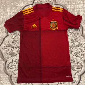Spaniens landslagströja Adidas röd - Röd fotbollströja från Adidas med Spaniens landslagsemblem på bröstet och gula detaljer. Klassisk V-ringning, korta ärmar och tre gula ränder på axlarna. Tillverkad i lätt funktionsmaterial (AEROREADY) som andas. Perfekt för match eller träning.