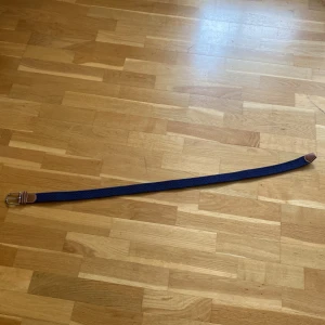 Mörkblå flätat bälte med skinn - Snyggt mörkblått flätat bälte med detaljer i brunt skinn och silvrigt spänne. Bältet har en stilren look med kontrastsömmar på skinnet och passar perfekt till jeans eller chinos. Den flätade strukturen gör det flexibelt och bekvämt att bära.