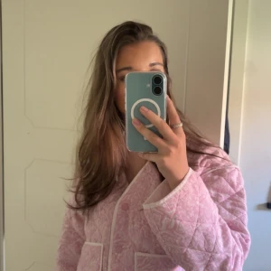 Quiltad rosa jacka med mönster - Säljer en quiltad rosa jacka med vitt paisleymönster och vita detaljer vid kanterna. Jackan har knappar framtill och två fickor. Perfekt för dig som vill ha en mysig och trendig look.